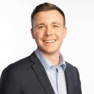 Headshot of Ben Jackson. Ben is a young white man with short brown hair. He is wearing a blue suit jacket with a blue dress shirt and no tie, standing in front of a white background. He is angled slightly away from the camera with his body and smiling.