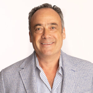 Headshot of Andrew Tow on a white background. Andrew is a middle-aged white man wearing a light gray sport coat with a blue checkered collared shirt underneath. He is smiling and facing the camera directly.