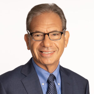 Headshot of Scott Schneider on a white background. Scott is a middle-aged white man, wearing glasses and a blue suit with a blue collared shirt and checkered tie. He is smiling and facing the camera.